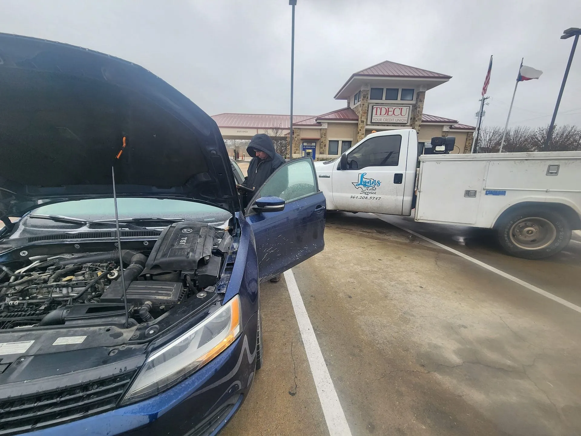 Person beside a blue car with the hood open, tow truck behind in a parking lot.