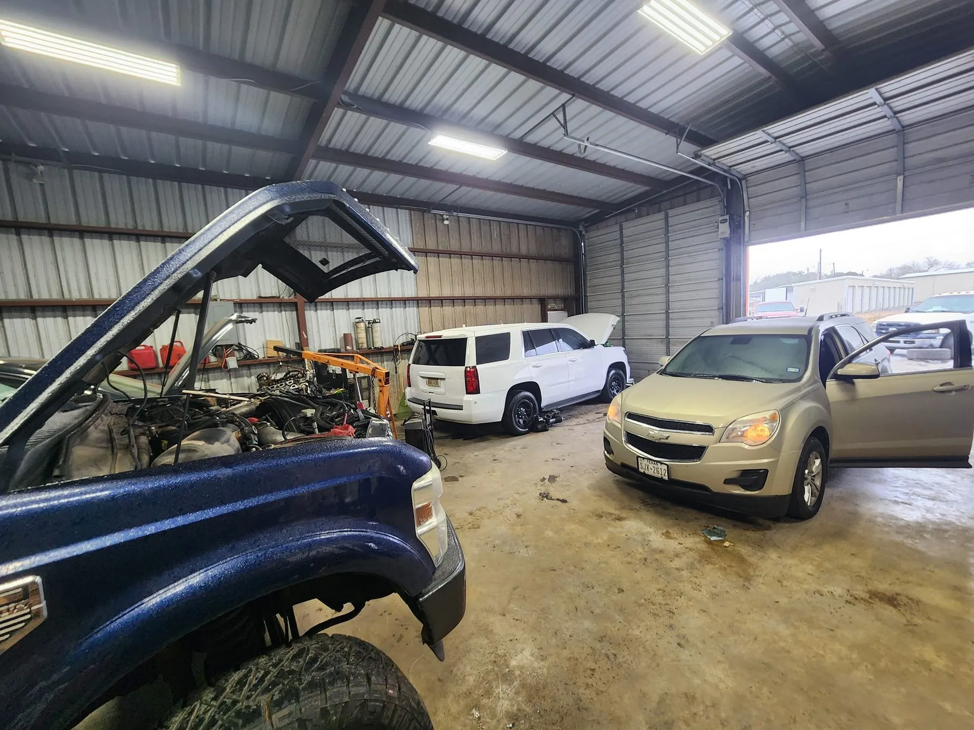 Cars in a garage with hoods open in a mechanic's workspace with tools and equipment.